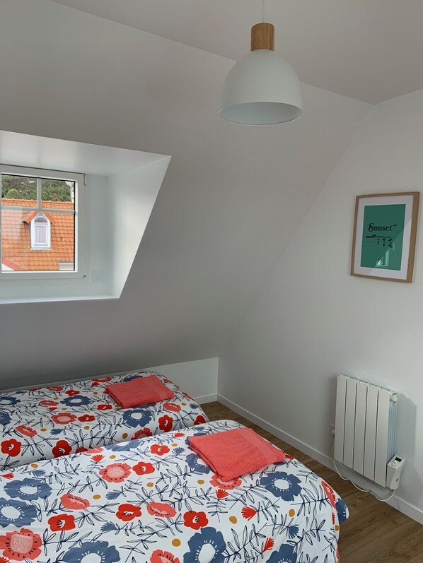 Un dormitorio con techo inclinado, una ventana con vista a un edificio de ladrillo, una cama con colcha estampada, un radiador blanco y un cuadro en la pared.