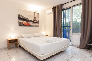 A hotel room with a bed, a nightstand, a wall-mounted lamp, a window with curtains, and a painting of the Eiffel Tower.