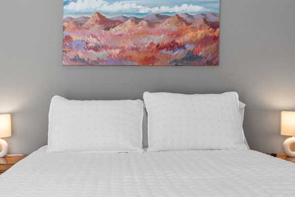 Una cama bien hecha con ropa de cama y almohadas blancas, un cuadro de montañas en la pared y dos lámparas de noche sobre mesitas de madera.