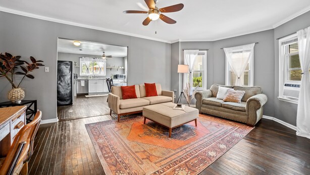Un salon spacieux avec un canapé beige, un pouf assorti et un grand tapis. On aperçoit un ventilateur de plafond, et une cuisine se trouve au fond de la pièce.