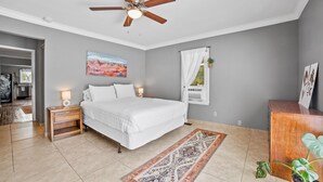 Une chambre à coucher comprenant un lit, des tables de chevet, un ventilateur de plafond, un tableau, une fenêtre avec des rideaux et une plante en pot.