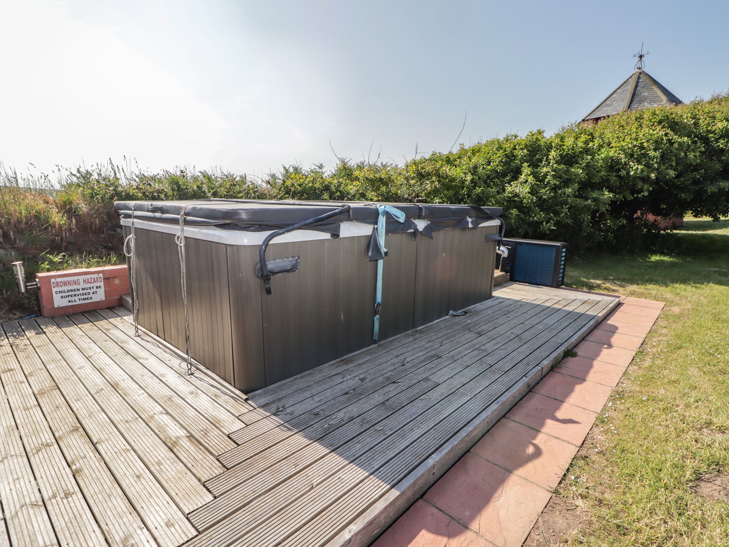A hot tub on a wooden deck with a safety sign, surrounded by greenery and a building in the background.