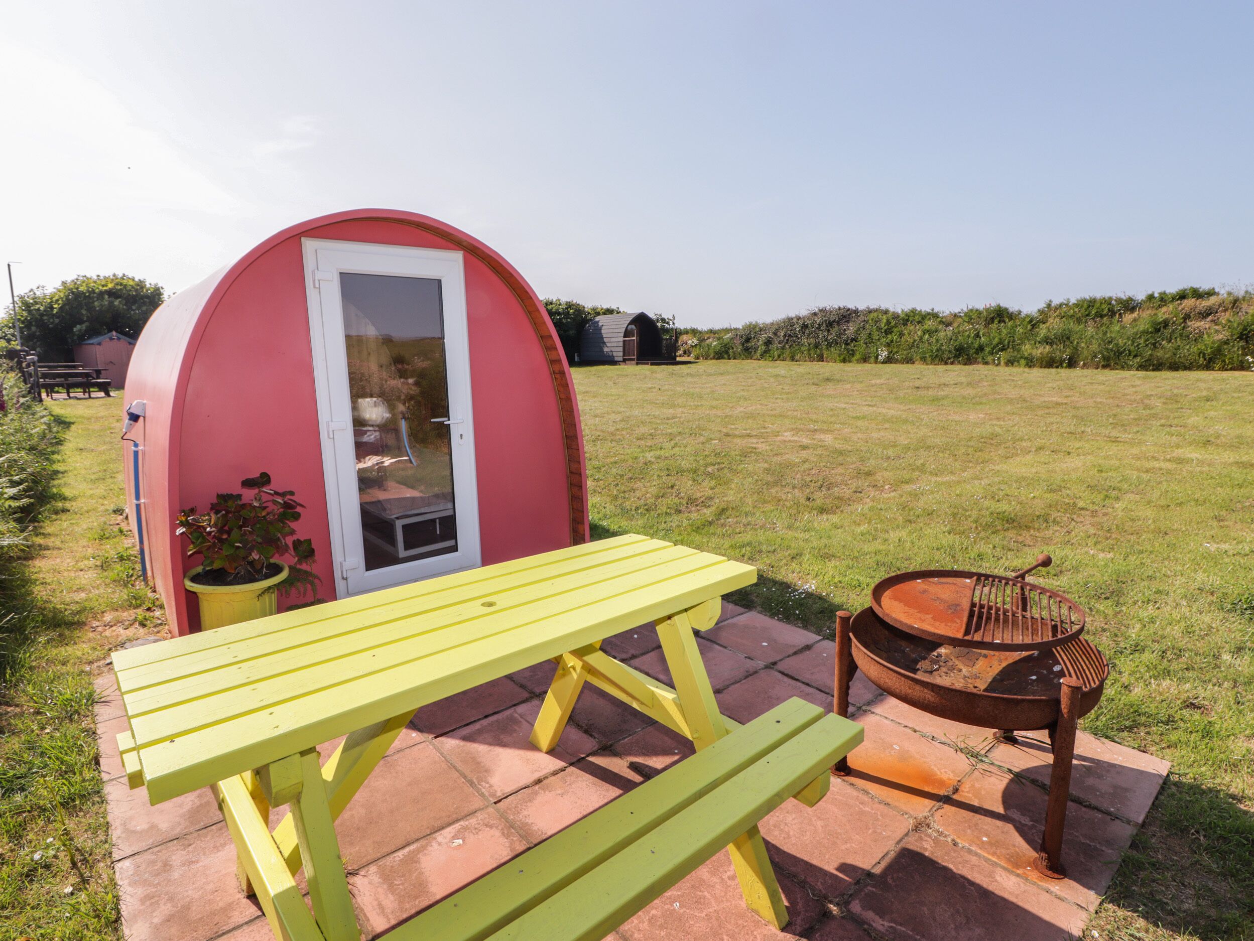 A small, pink, semi-circular structure with a door and windows, a yellow picnic table, and a metal chair.