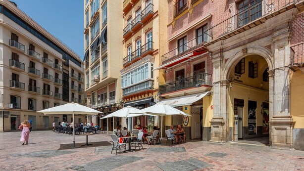 Un área de café al aire libre con mesas y sillas bajo sombrillas, rodeada de edificios de varios pisos con balcones y tiendas.