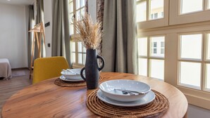 Una mesa de comedor con platos y cubiertos, un florero con plantas secas, y una silla amarilla en una habitación con ventanas grandes y cortinas.