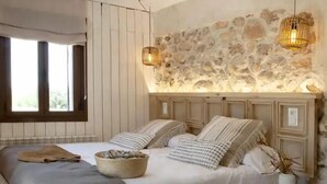 Ein Schlafzimmer mit Steinwand, Holz-Kopfteil, zwei Nachttischlampen, einem Bett mit Kissen und Decke sowie einem Nachttisch mit Vase und einem Fenster mit Vorhängen.