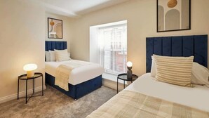 A hotel room with two beds, a window with blinds, a bedside table with a lamp, and a framed picture on the wall. - Host & Stay | 3 The Mount (Whitby)
