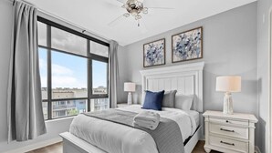 Un dormitorio con un ventanal grande, una cama con ropa de cama blanca, una mesita de noche con lámpara y dos cuadros colgados en la pared.