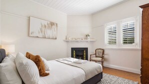 A bedroom with a bed, a fireplace, a chair, and a window with shutters. - Sampson Cottage | Historic Charm (Orange)