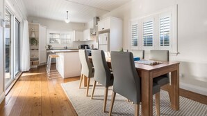 A modern kitchen with white cabinets, a dining area with a wooden table and chairs, and a large window with blinds. - Sampson Cottage | Historic Charm (Orange)