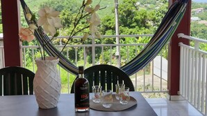 Une table avec une bouteille de spiritueux, des verres et un vase de fleurs, le tout placé devant un paysage pittoresque, visible à travers un hamac.