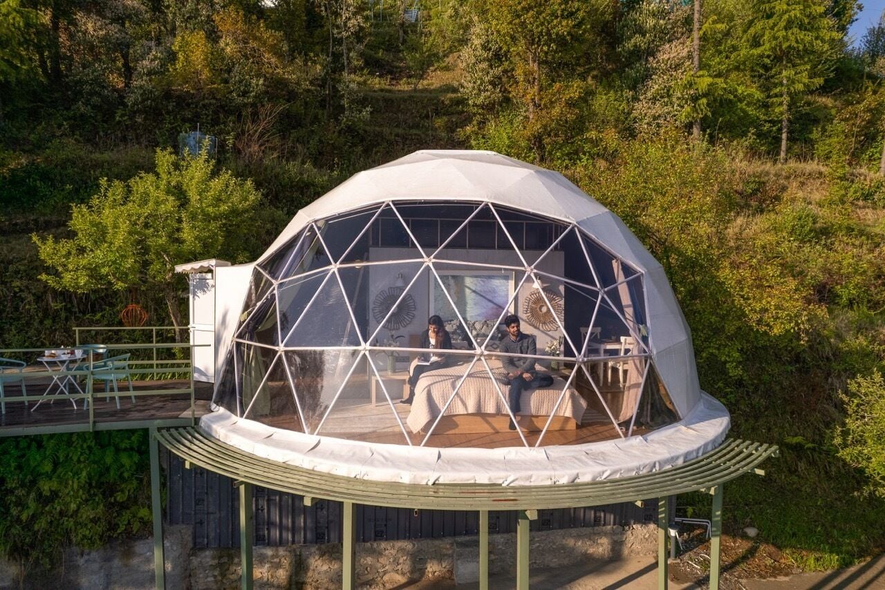 A geodesic dome structure with a transparent roof, a wooden deck, and a view of a forested hillside.