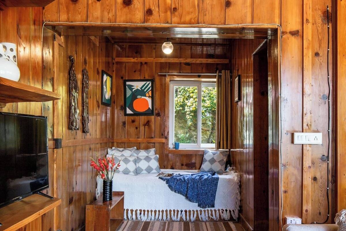 A cozy cabin interior with a wooden interior, a bed with a blue blanket, a television, and a window with a view of greenery.