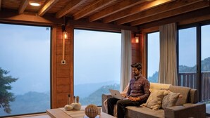 A person sitting on a sofa with a laptop, a wooden table with decorative items, and large windows with a view of a mountainous landscape. - Avalon Cottages Kanatal (Tehri)