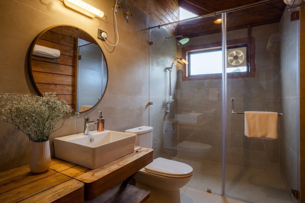 Une salle de bain avec un meuble-lavabo en bois, un miroir rond, un lavabo blanc, des toilettes et une douche séparée par une paroi en verre.