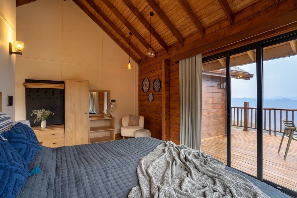 A bedroom with a large bed, wooden ceiling, a desk, a chair, and a view of a balcony with a wooden deck and mountains in the background.