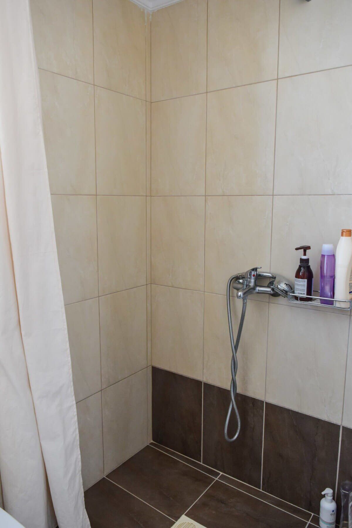 A shower area with beige and dark brown tiles, a showerhead, and a shelf with toiletries.