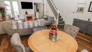 Un salon avec une table ronde en bois, des fauteuils à revêtement gris, un canapé gris agrémenté d’un plaid en fourrure, une cheminée et un escalier.