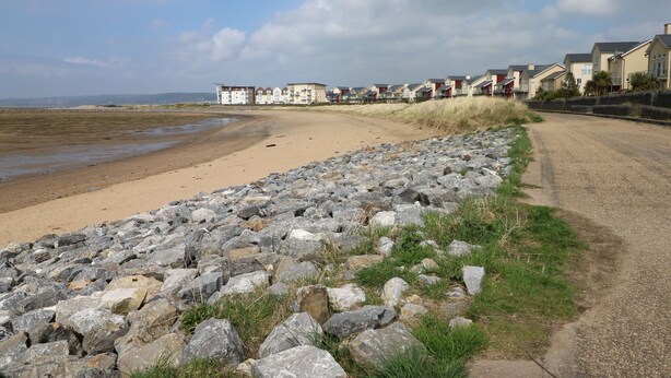 Une route côtière bordée d’une plage de sable et d’une côte rocheuse. Des maisons sont construites le long de la route, et la plage semble être restée à l’état sauvage.