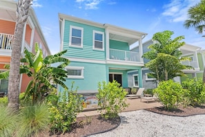 A two-story building with a balcony, surrounded by tropical plants and a gravel driveway.