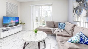 Ein modernes Wohnzimmer mit einer Couch, einem Couchtisch, einem Fernseher auf einer weißen Unterhaltungsanlage und einem großen Fenster mit Jalousien.