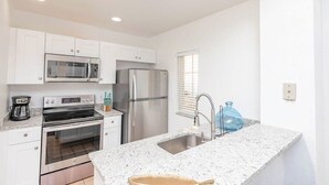Una cocina moderna con armarios blancos, electrodomésticos de acero inoxidable, encimera de granito y una ventana con persianas.