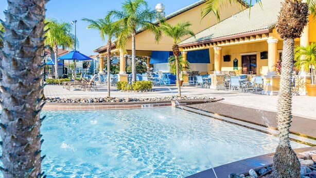 Zona de piscina con sillas de descanso, palmeras y un edificio con tejado de azulejos.