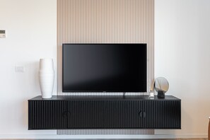 A modern living room with a large flat-screen TV on a black TV stand, a white vase, and a small round mirror. - Magical  2 BDR/Parking By Isrentals (Jerusalem)