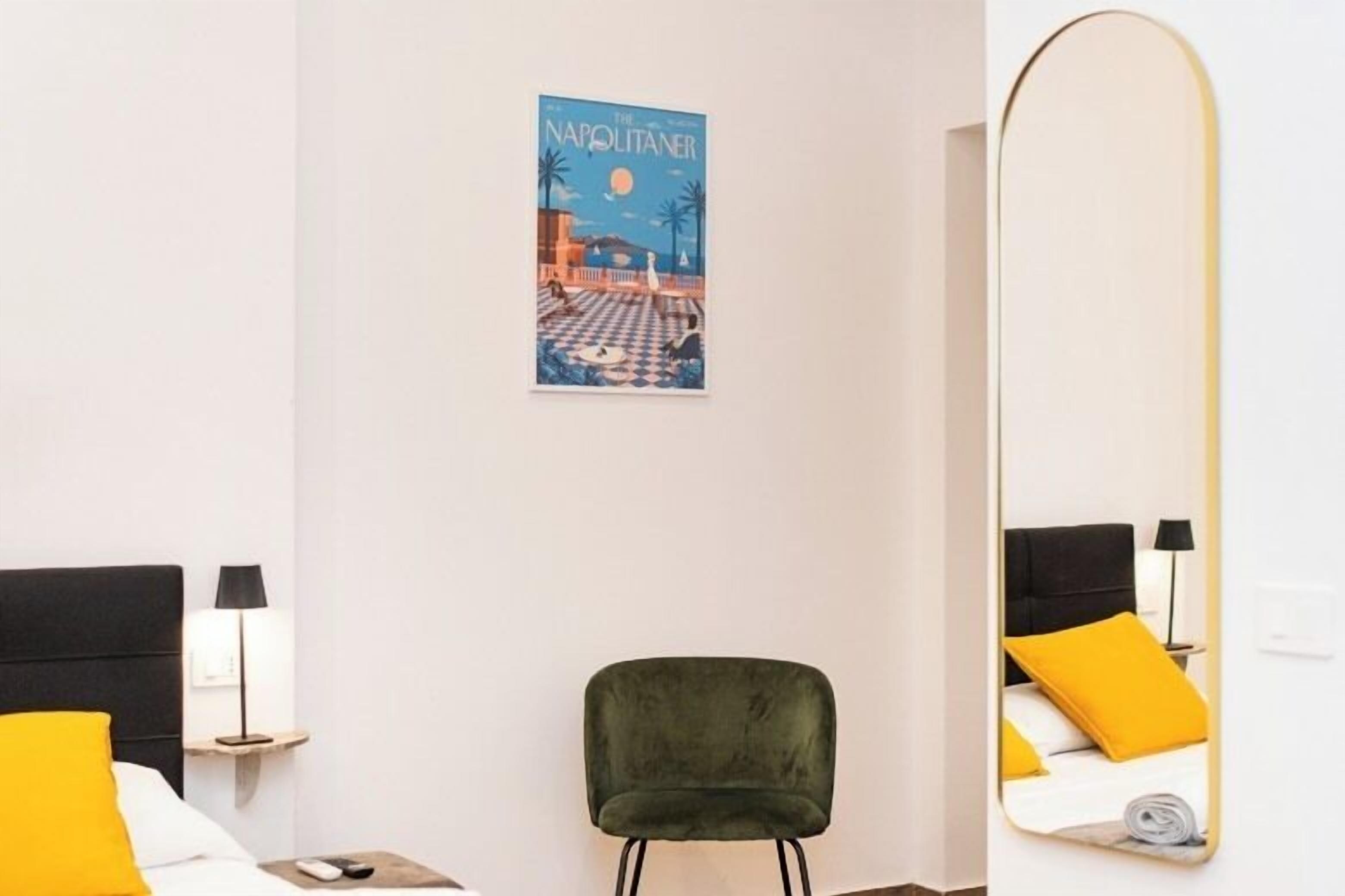 Quarto de hotel com cama, cadeira, espelho, pôster fixado na parede e mesa de cabeceira com abajur.
