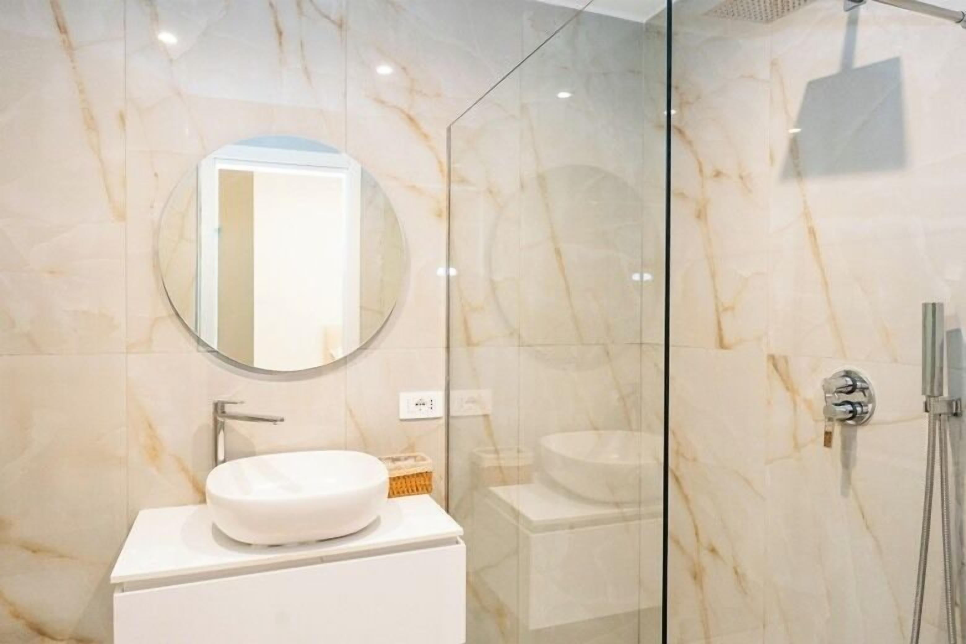 Une salle de bain moderne avec un grand miroir rond, un lavabo blanc et une douche séparée par des parois en verre.