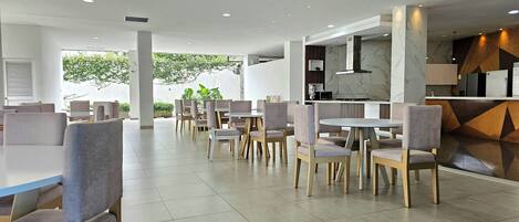 A modern restaurant with a clean, minimalist interior, featuring grey upholstered chairs and white tables, a marble backsplash, and a view of greenery outside.