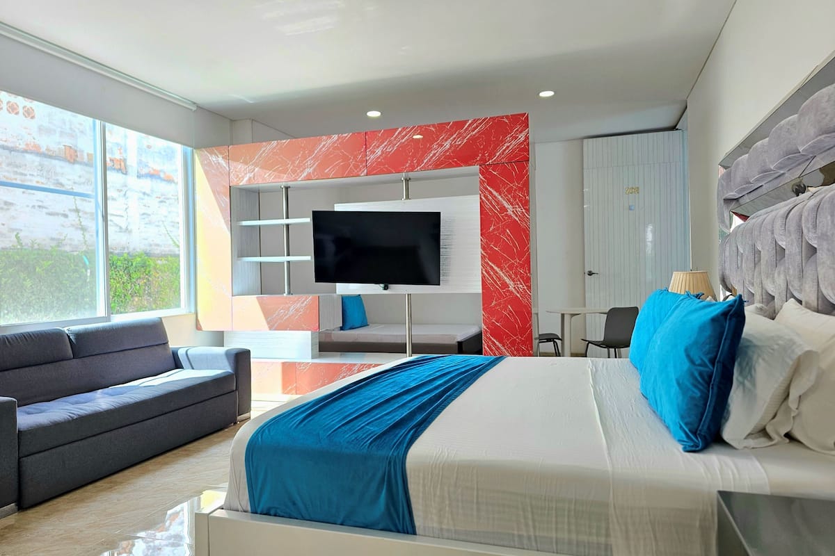 Een moderne hotelkamer met een groot bed, een bank, een televisie bevestigd aan een rode en witte marmeren muur, en een raam met uitzicht op groen.