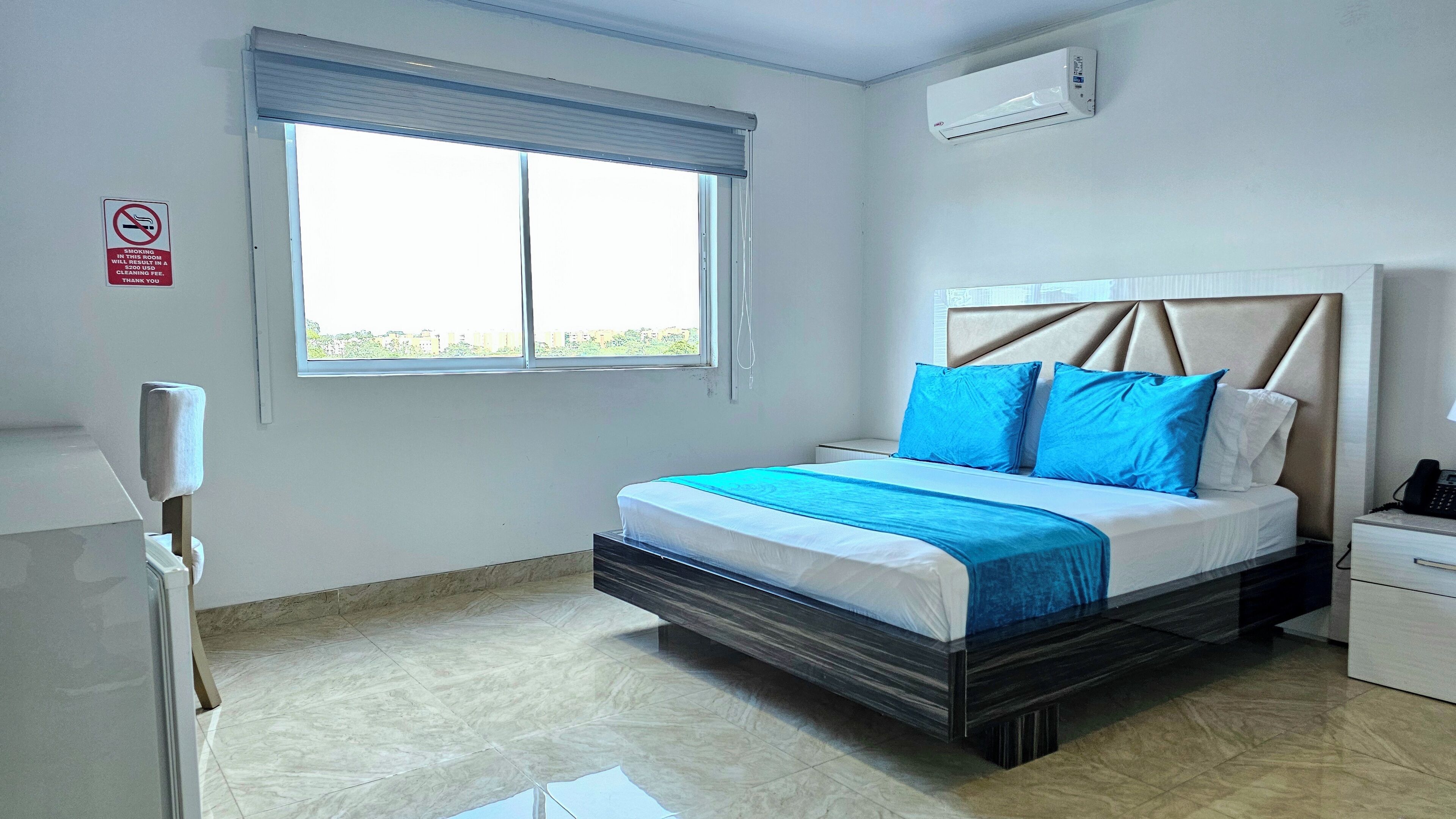 A hotel room with a bed, a window with blinds, an air conditioner, and a phone on a bedside table.