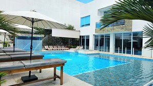 A swimming pool with lounge chairs and umbrellas, surrounded by a modern building with large windows. - Lotto Hotel Boutique (Cali)