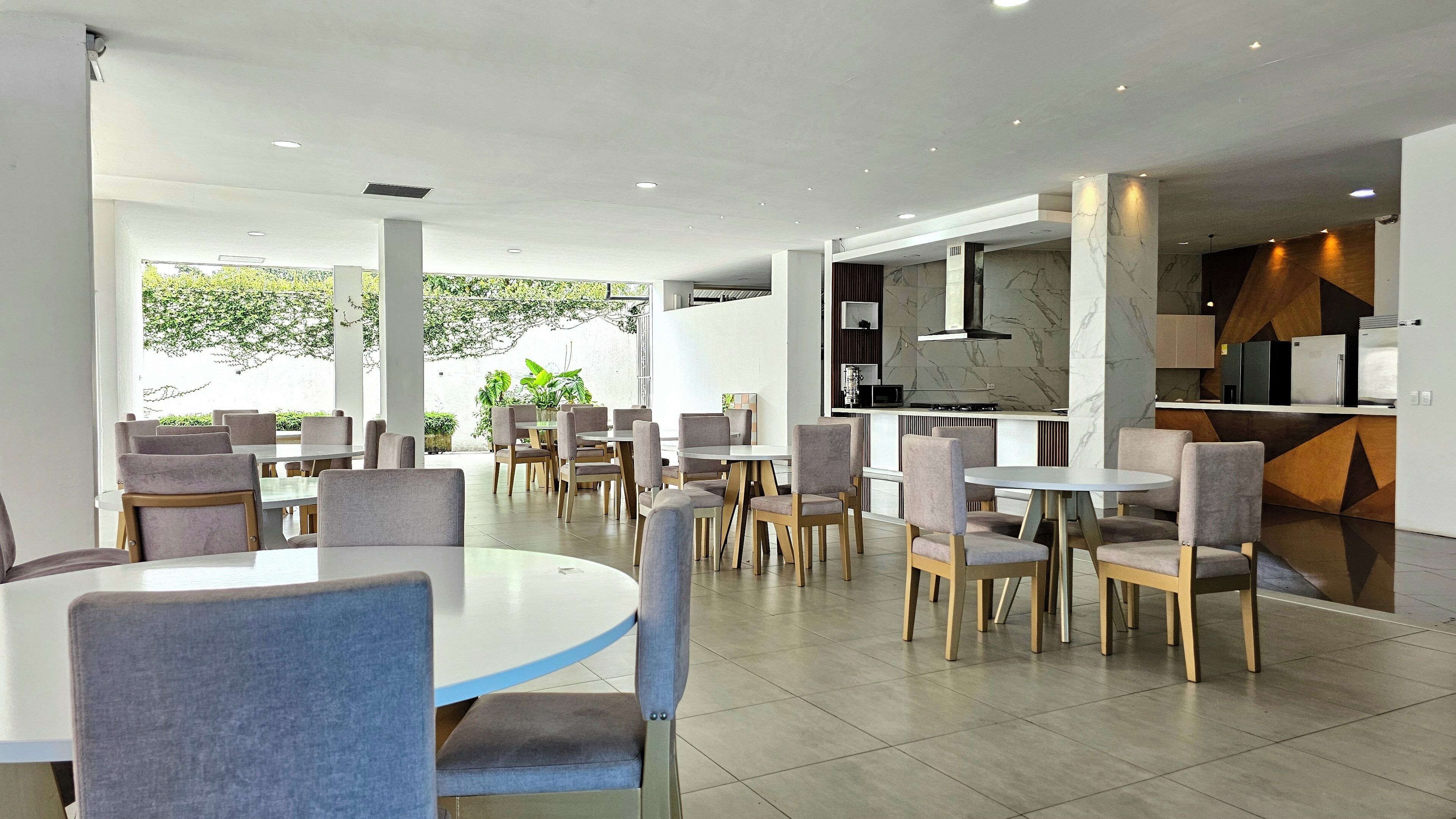 A modern dining area with round tables and chairs, a kitchen with marble countertops, and a view of greenery outside.