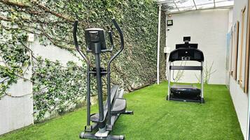 A small outdoor gym area with an elliptical machine and a treadmill, surrounded by a white wall and a green artificial grass floor.