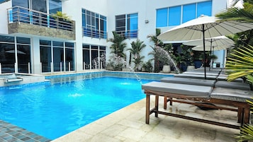 Una piscina con sillones de descanso y sombrillas, rodeada por un edificio con amplias ventanas y un balcón.