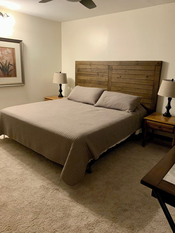 Una cama bien hecha con cabecera de madera, dos mesitas de noche con lámparas, un cuadro en la pared y suelo cubierto de alfombra.