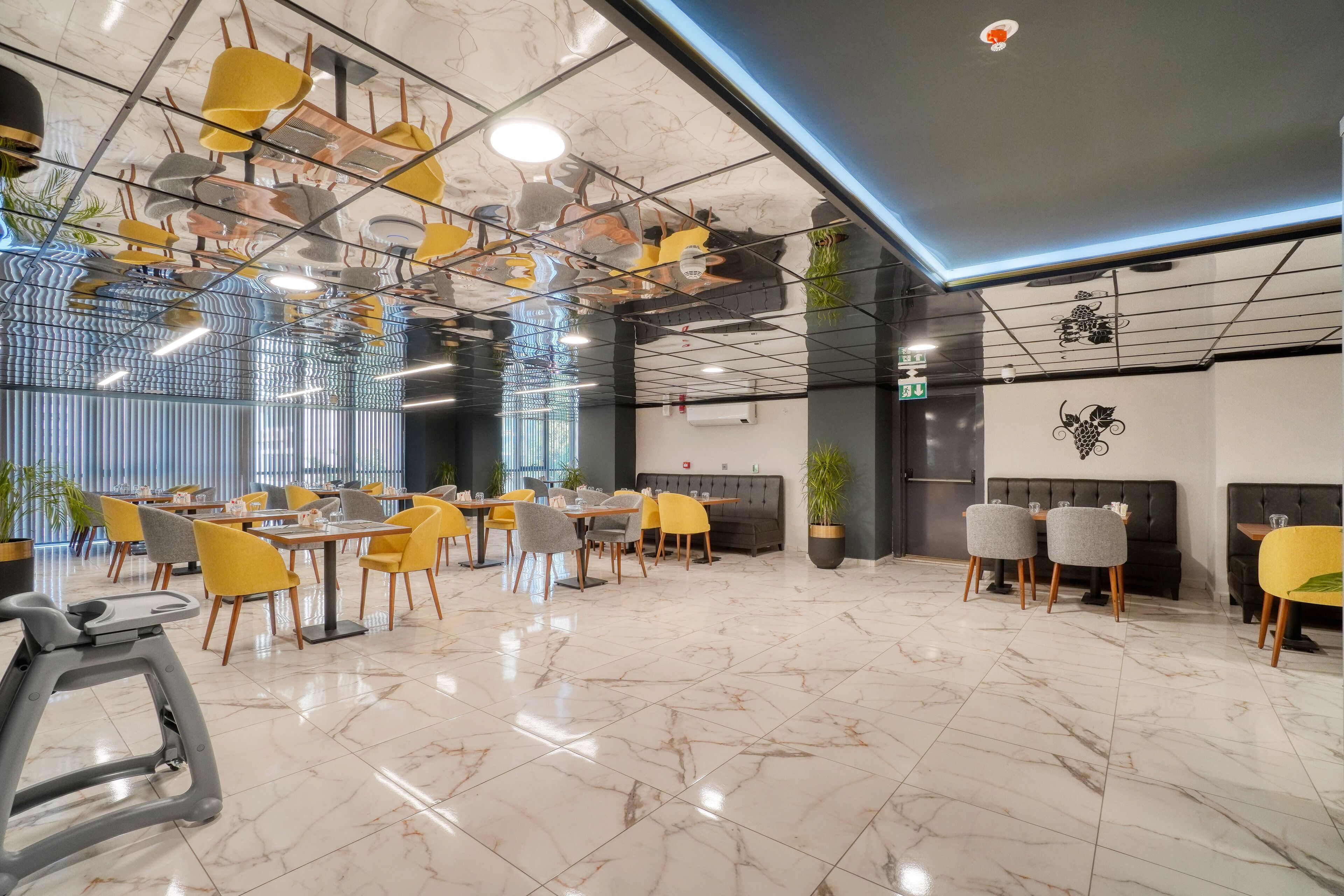 A modern restaurant with a high ceiling, marble flooring, and a mix of yellow and grey chairs.