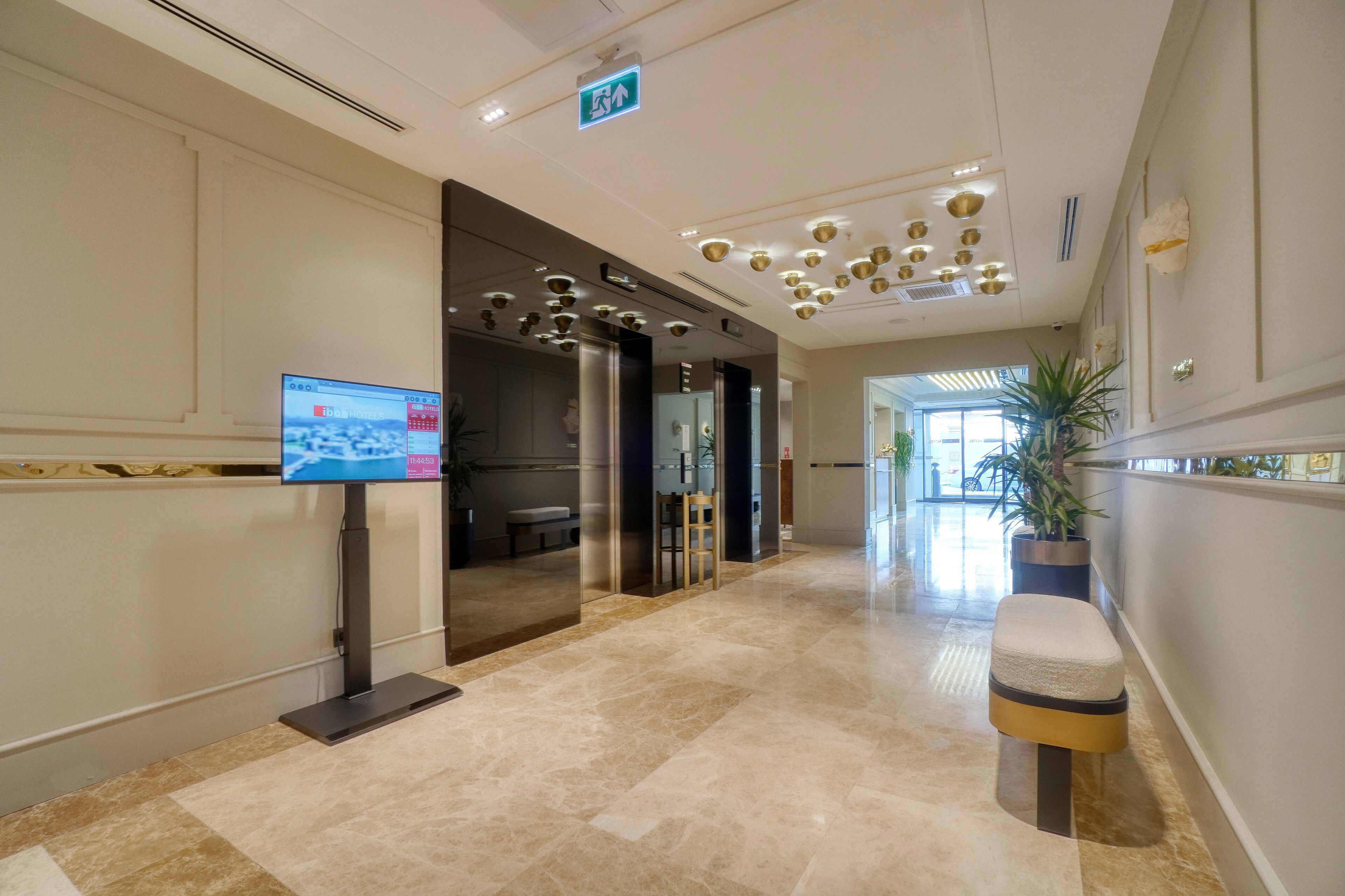 A modern hotel lobby with a marble floor, a reception desk, a television screen, and a potted plant.