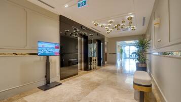 A modern hotel lobby with a marble floor, a reception desk, a television screen, and a potted plant.