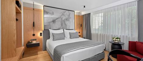 Un dormitorio moderno con una cama grande, mesitas de noche, una silla roja y vistas a la vegetación a través de cortinas traslúcidas.
