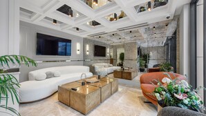 A modern living room with a white sofa, a glass coffee table, and a red armchair. There is a large flat-screen TV mounted on the wall and a stone wall in the background. - IBOS HOTELS IZMIR (Izmir)