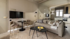 Un salon moderne avec un canapé, deux tables d’appoint, un téléviseur et, au fond, un coin cuisine.