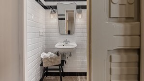 Une salle de bain avec des murs recouverts de carreaux blancs rectangulaires, un lavabo blanc et un miroir au-dessus. On y trouve un panier à serviettes sur le sol et une serviette accrochée au mur.