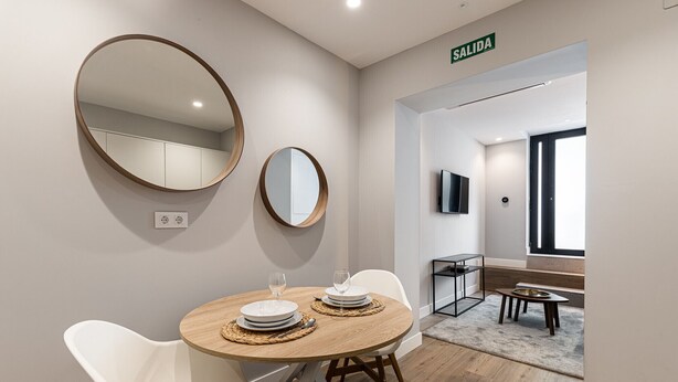 Ein modernes Hotelzimmer mit einem runden Esstisch, zwei weißen Stühlen und zwei runden Spiegeln an der Wand. Ein Schild weist auf den Ausgang, ein Fernseher ist an der Wand montiert und es gibt ein Fenster mit Aussicht.