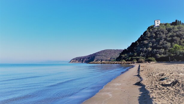 Une plage bordée d’une mer d’un bleu limpide, d’un rivage sablonneux et d’une colline surmontée d’un bâtiment.