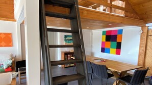 Een loft-achtige kamer met een houten plafond, een houten trap, een whiteboard met een kleurrijk schema, en een eethoek met een tafel en stoelen.