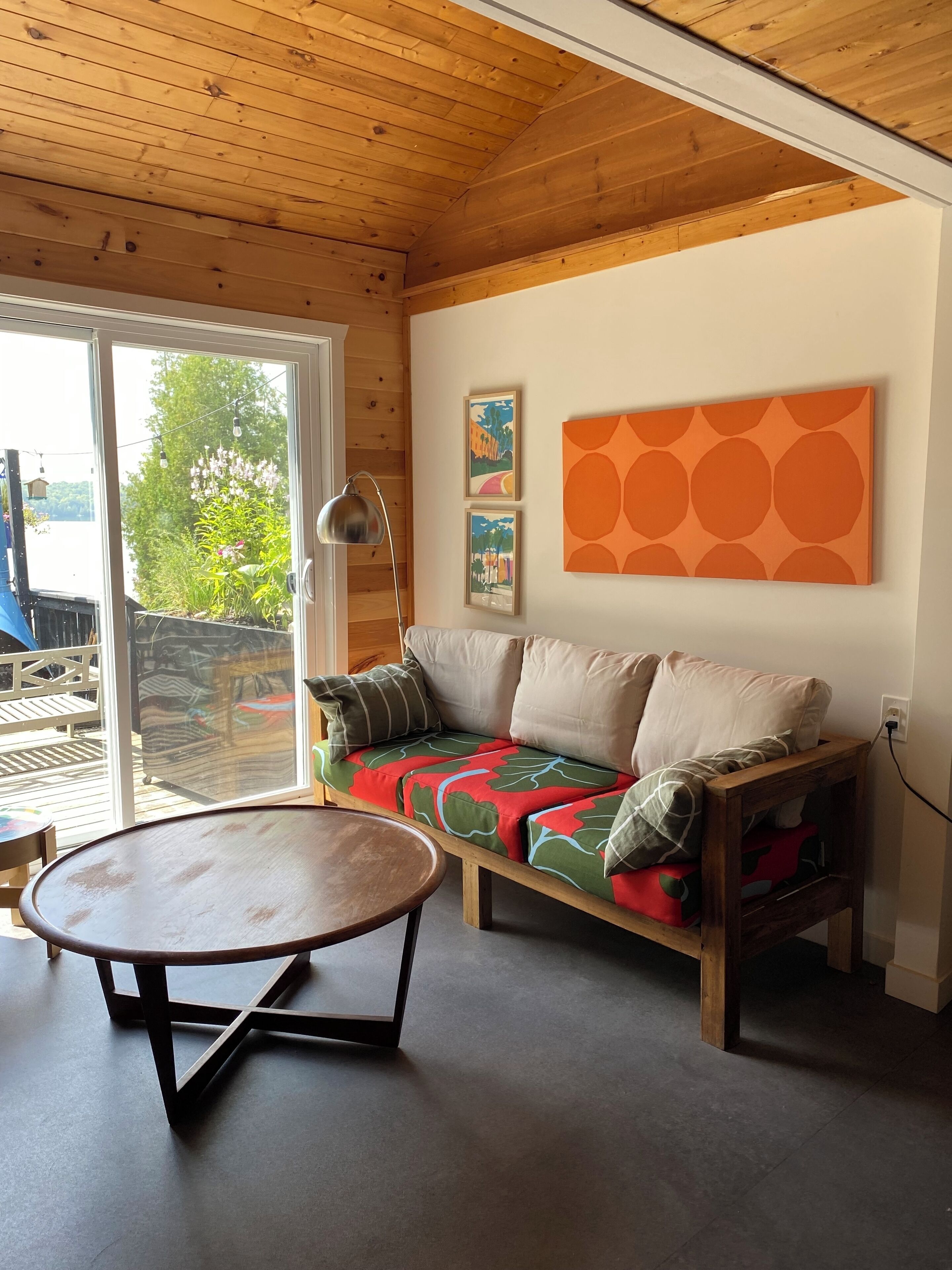 Een moderne woonkamer met een houten plafond, een bank met kleurrijke kussens, een ronde tafel en een groot raam met uitzicht op groen.
