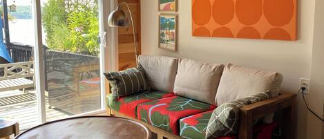 Een moderne woonkamer met een houten plafond, een bank met kleurrijke kussens, een ronde tafel en een groot raam met uitzicht op groen.